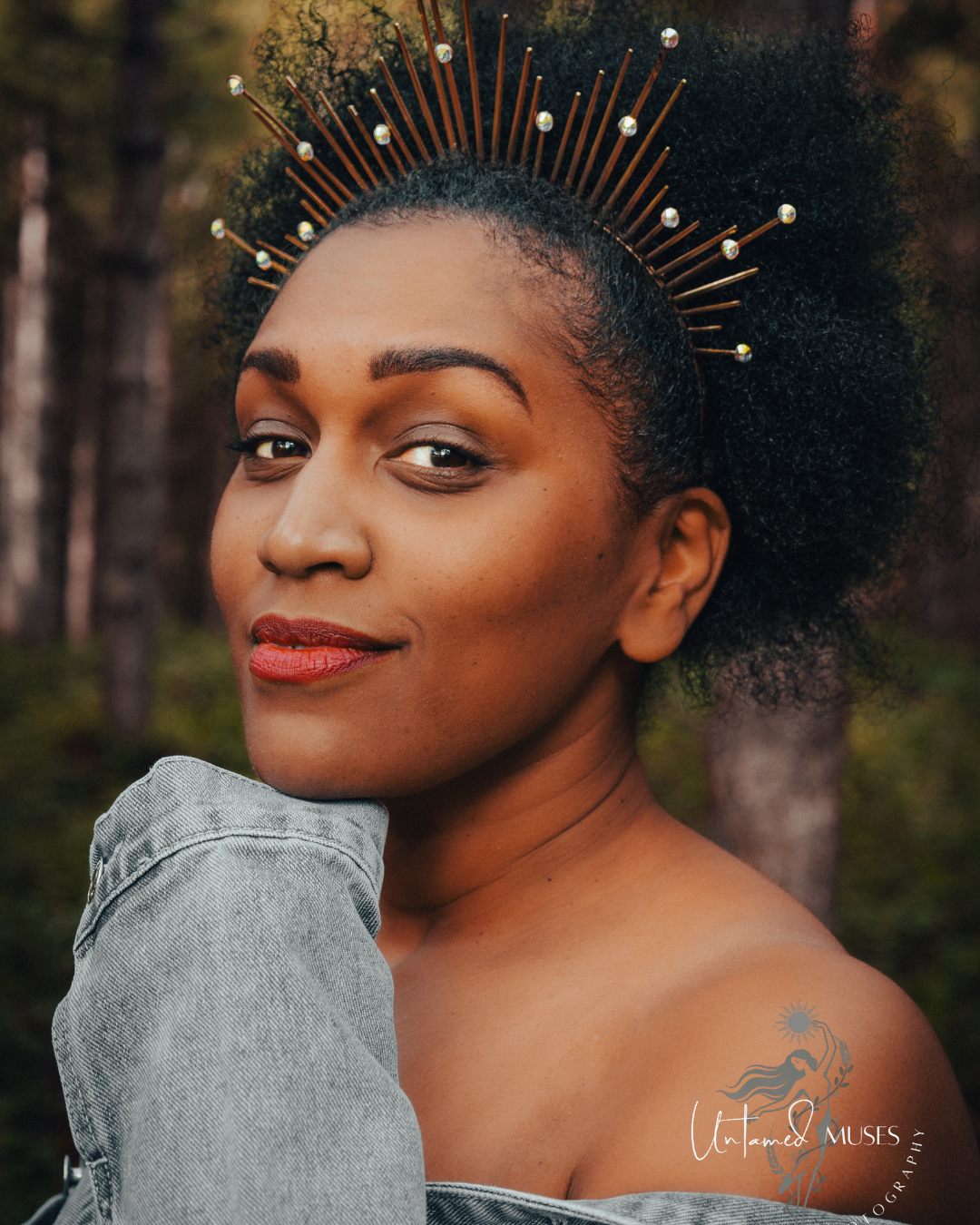A close-up portrait of a woman outdoors with dark curly afro hair styled in a high crown-like updo adorned with metallic spikes and small pearl-like beads. She is wearing a light blue denim off-the-shoulder top, with a blurred forest background.