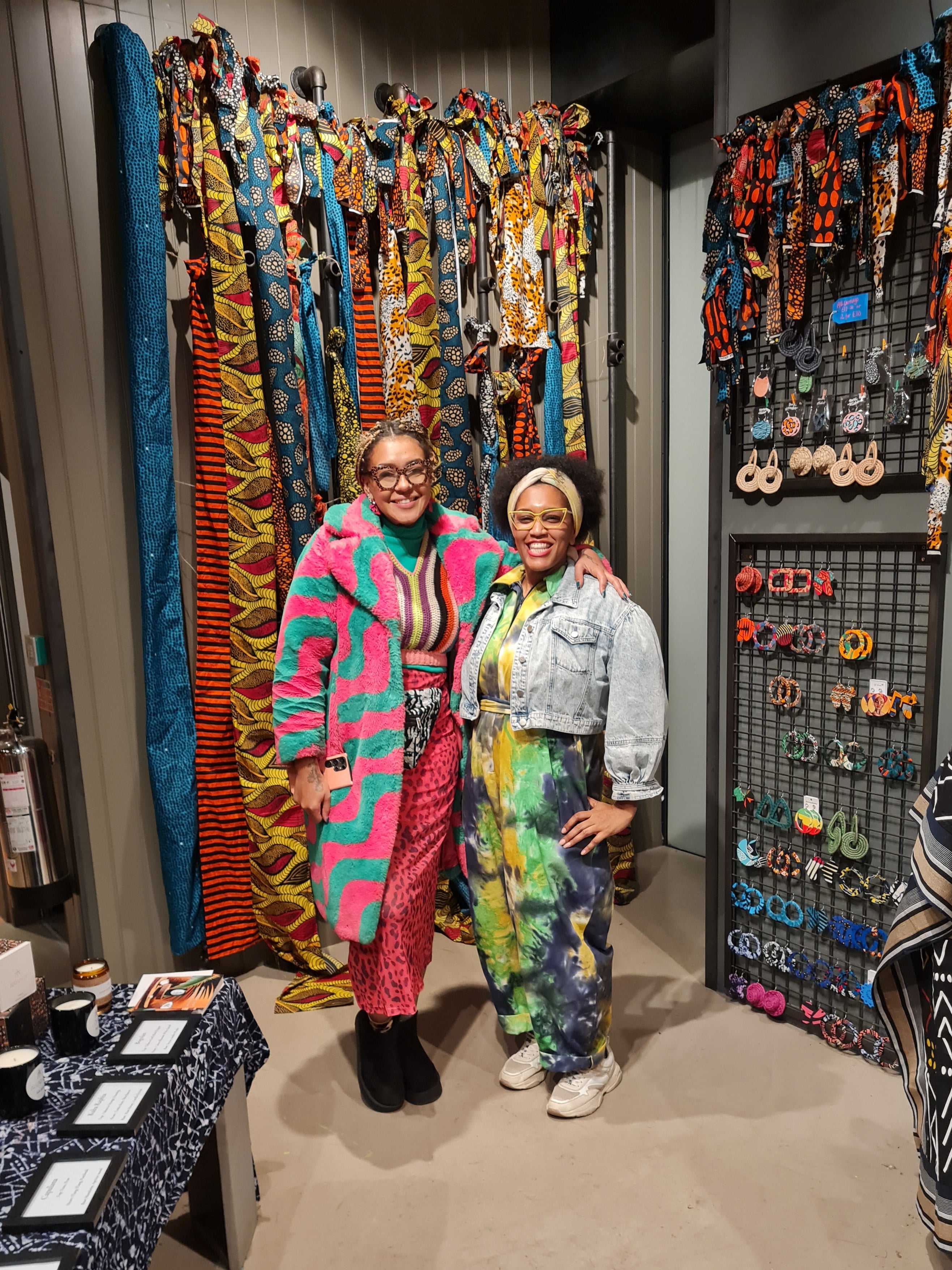 Two women posing together in a vibrant, stylish boutique interior. The woman on the left wears a bright pink and teal patterned coat over a red dress, while the woman on the right wears a light denim jacket and a colorful tie-dye jumpsuit.