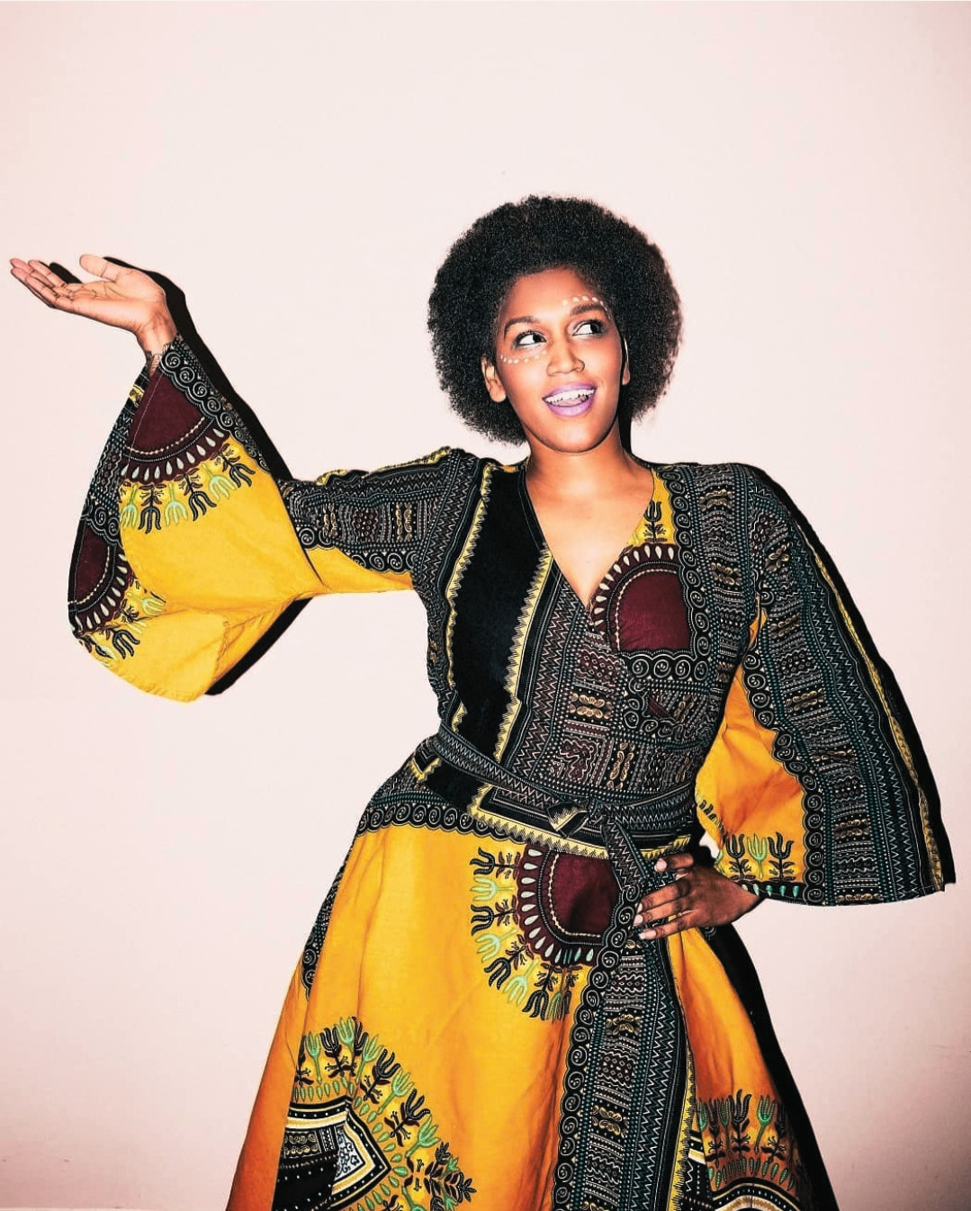 A woman modeling a vibrant yellow and black African wax print kaftan dress with wide bell sleeves and a matching belt, posed against a solid pale pink background.
