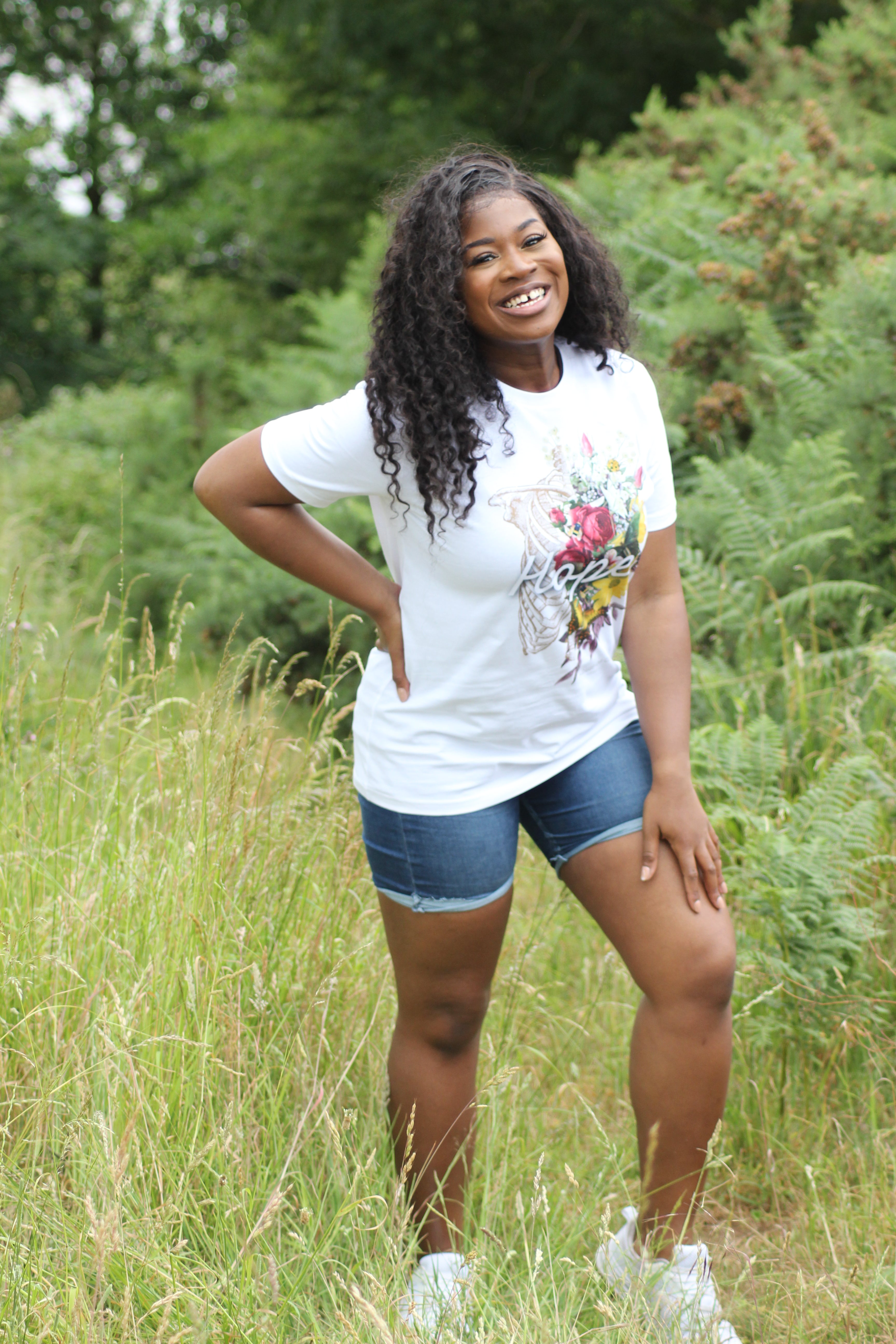 Young black woman standing in a grassy field wearing a white t-shirt with a floral design.