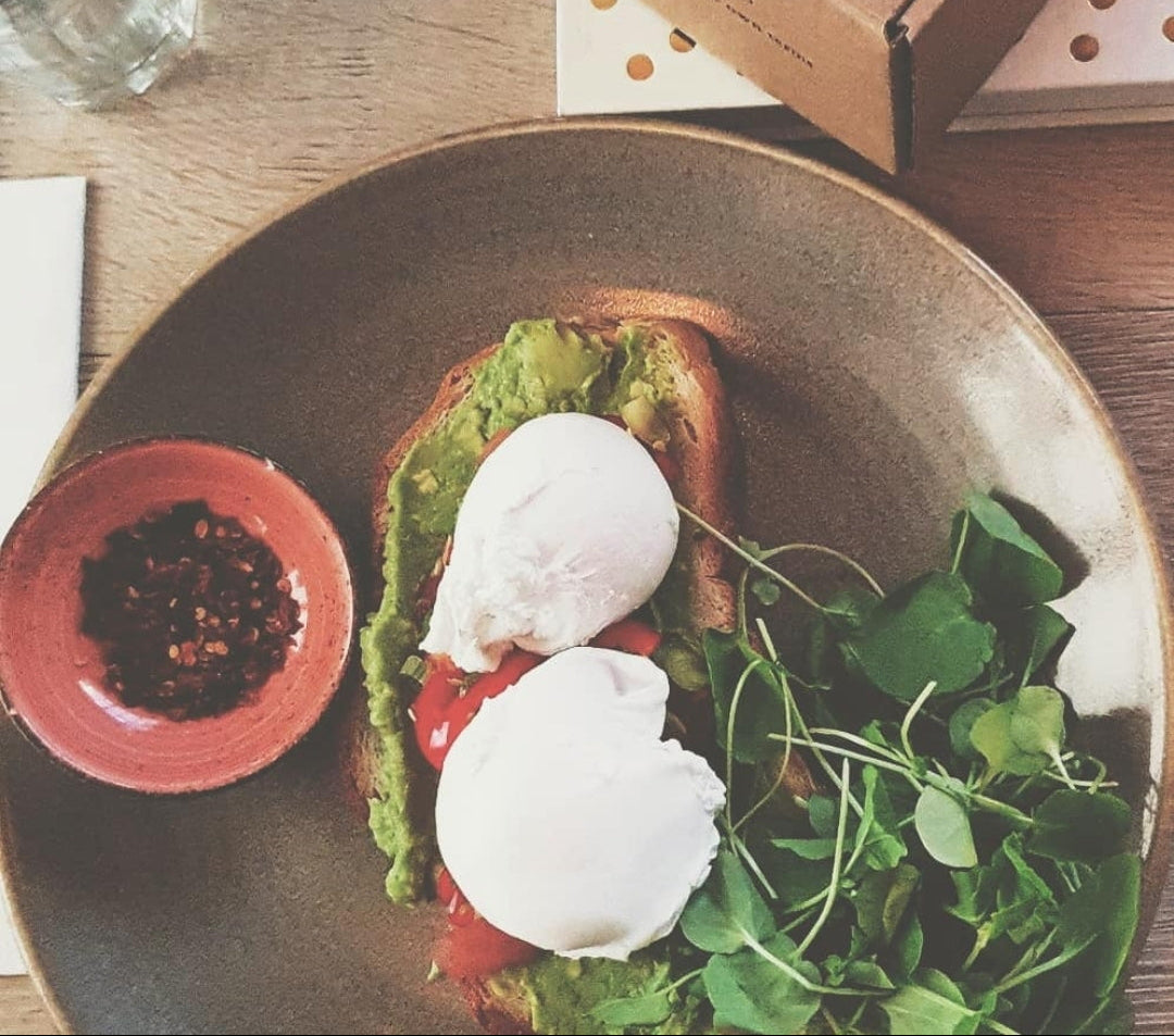 An Instagram-style overhead photo shows a plated avocado toast with two poached eggs, greens, and a small bowl of red chili flakes on a wooden table, alongside a brown gift box .