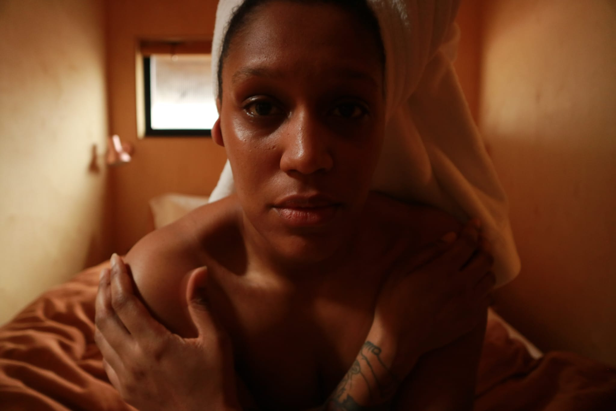 A close-up lifestyle shot of a woman with a white towel wrapped around her head, sitting on bed with warm orange-toned lighting. Her face is blurred, sad, disconnected and she has a tattoo on her left arm.