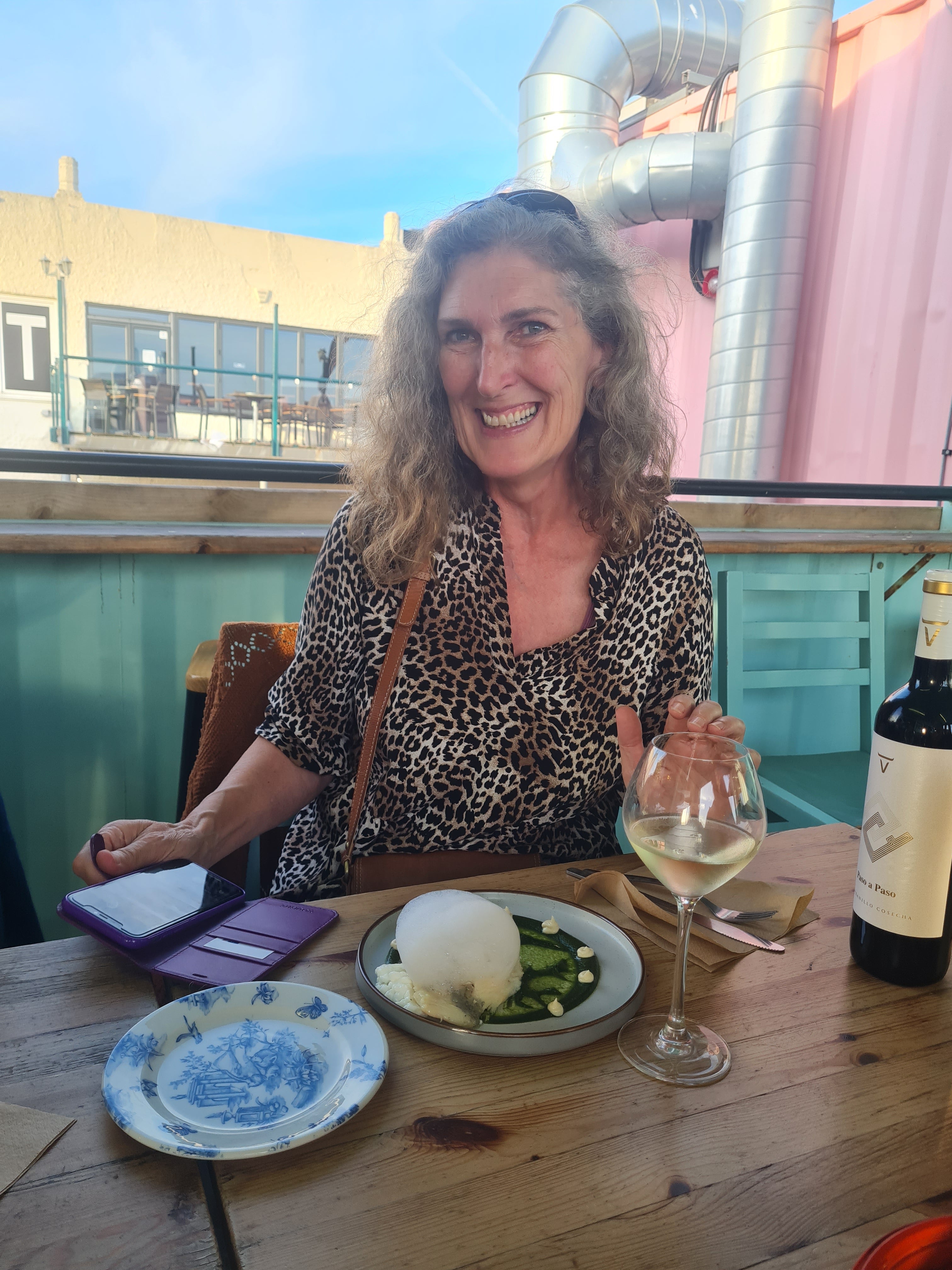 A smiling woman with blonde curly hair sits at a wooden table on an outdoor patio, wearing a leopard-print top. In front of her are a plate of food, a glass of white wine, a purple wallet, and a wine bottle.