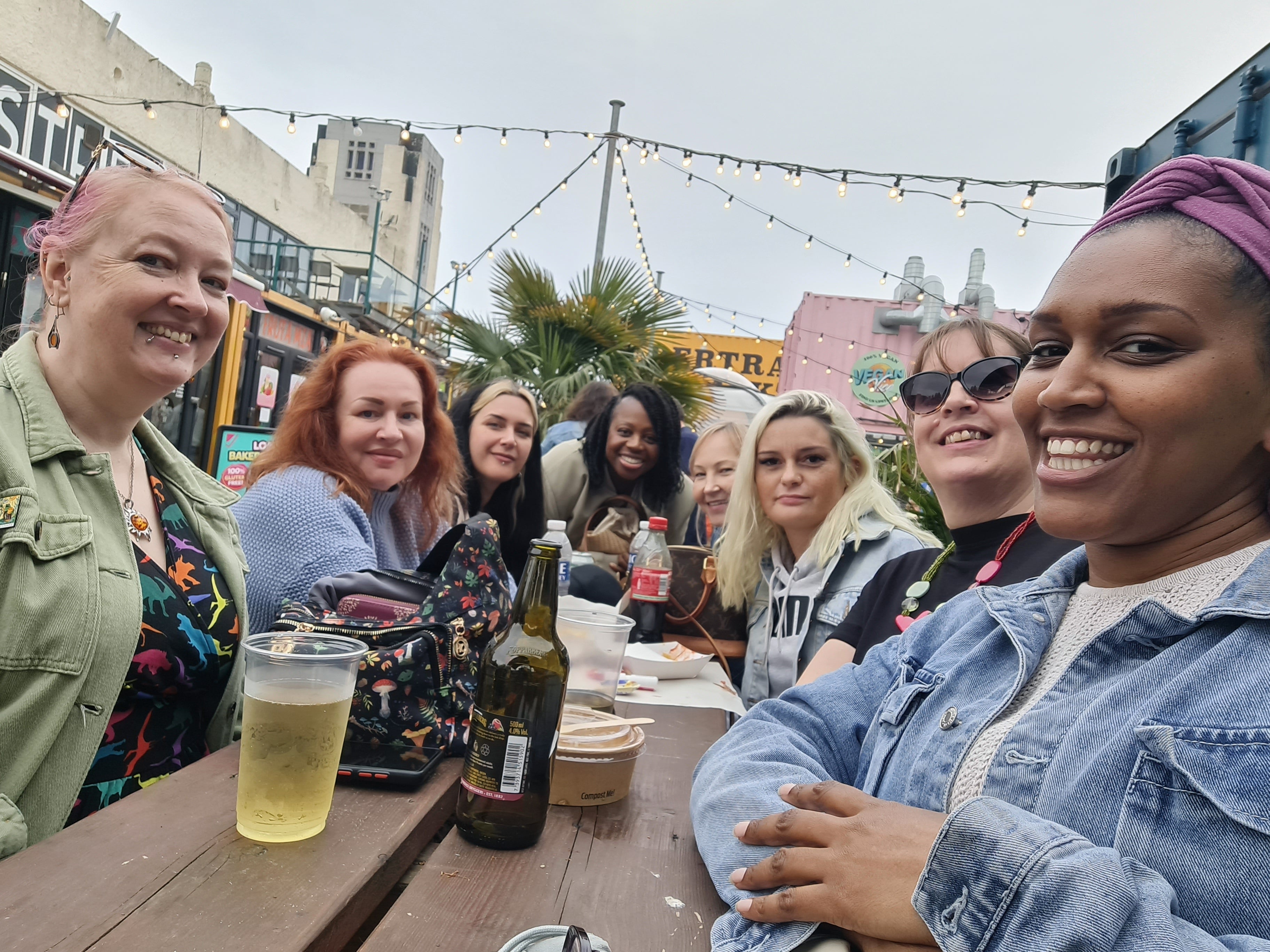 A group of seven women smiling and laughing together at an outdoor wooden table, with beer bottles and cups on the table. The setting is a lively urban street patio with buildings, string lights, and a cloudy sky in the background.
