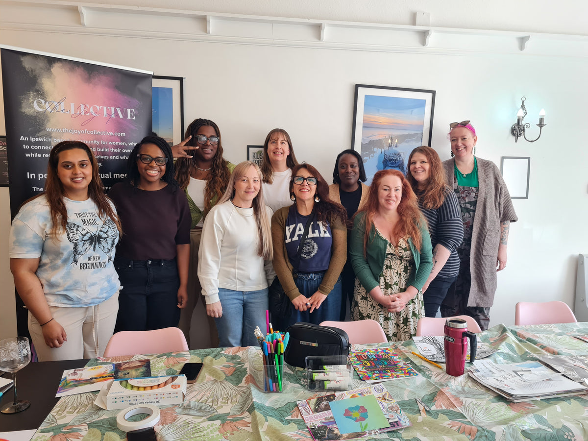 A group of ten women posing together in a bright indoor studio or workshop space, standing behind a decorated table with art supplies, magazines, and a colourful banner that reads 'The Joy of Collective'.
