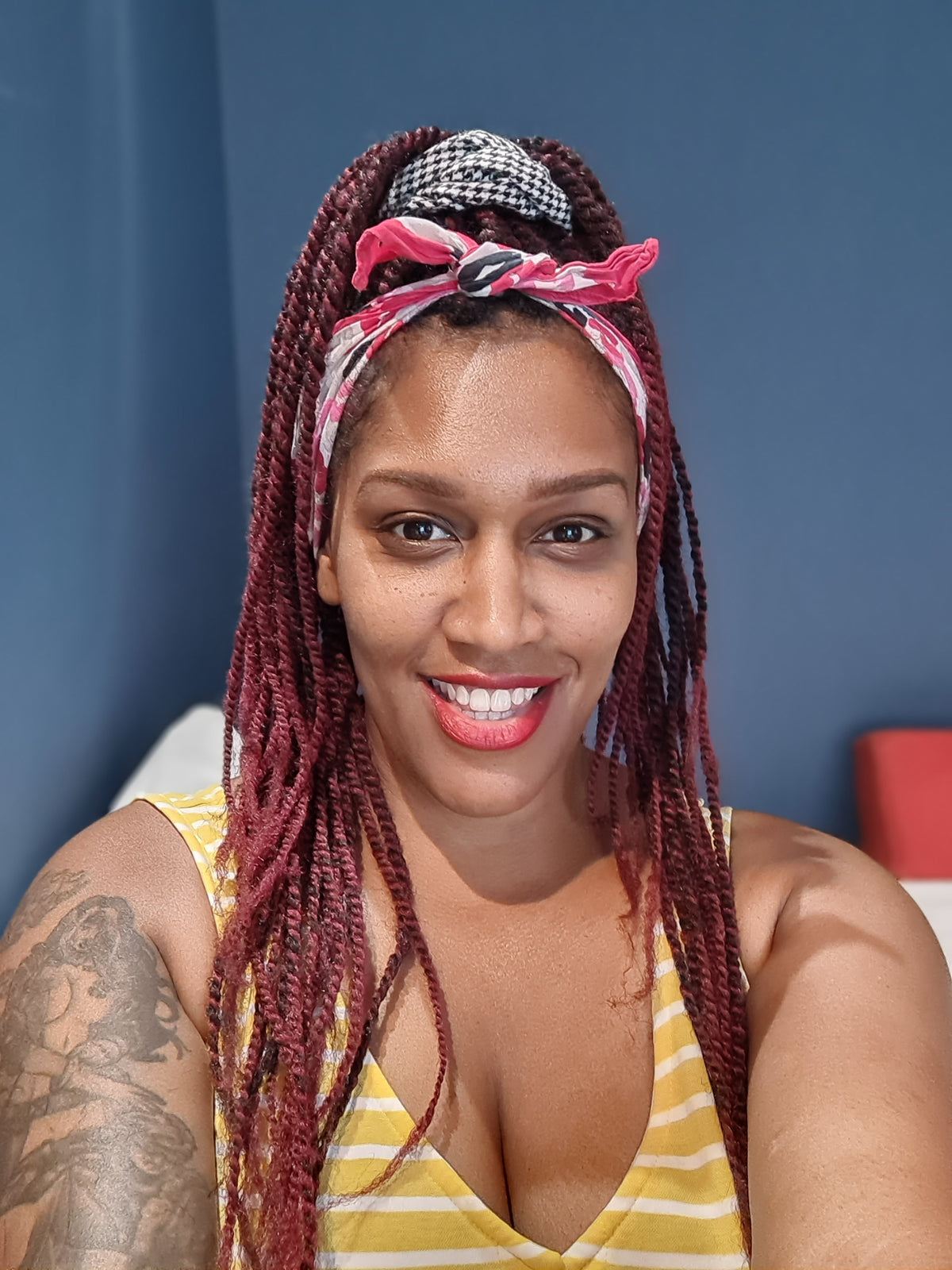 A woman with long burgundy braided hair wearing a pink and white patterned headband, a black and white houndstooth hair tie, and a yellow and white striped tank top, posing against a blue wall.