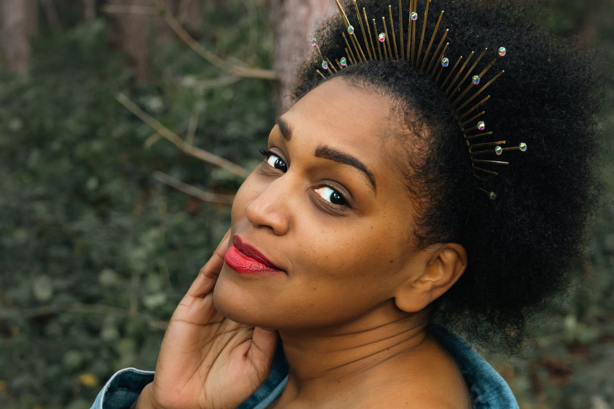 A close-up portrait of a black woman with dark curly afro hair wearing a gold spiked tiara adorned with small colourful rhinestones, set against a blurred forest background.