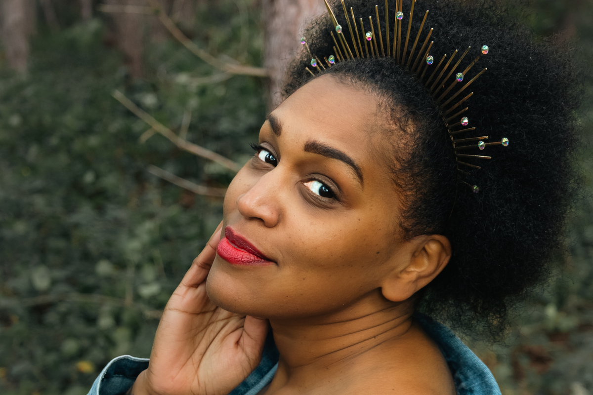 A close-up portrait of a black woman with dark curly afro hair wearing a gold spiked tiara adorned with small colourful rhinestones, set against a blurred forest background.