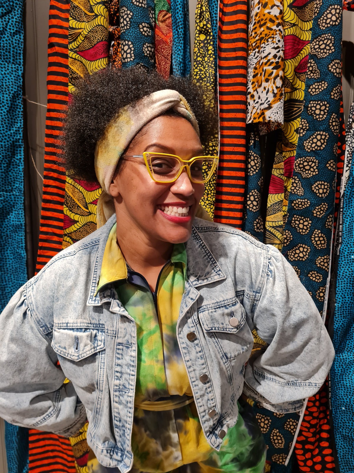 A smiling woman with an afro wearing a light-wash denim jacket over a green and yellow tie-dye shirt, standing in front of a colorful display of patterned fabrics.